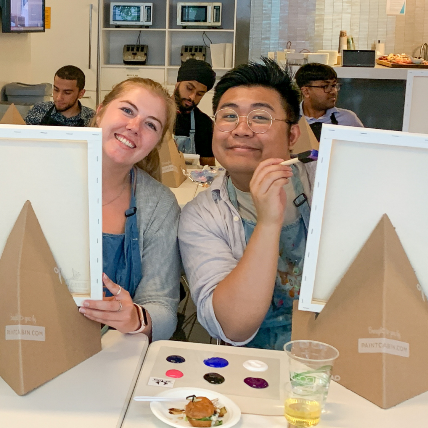 Two participants painting and smiling during an offsite team building activity with Paint Cabin